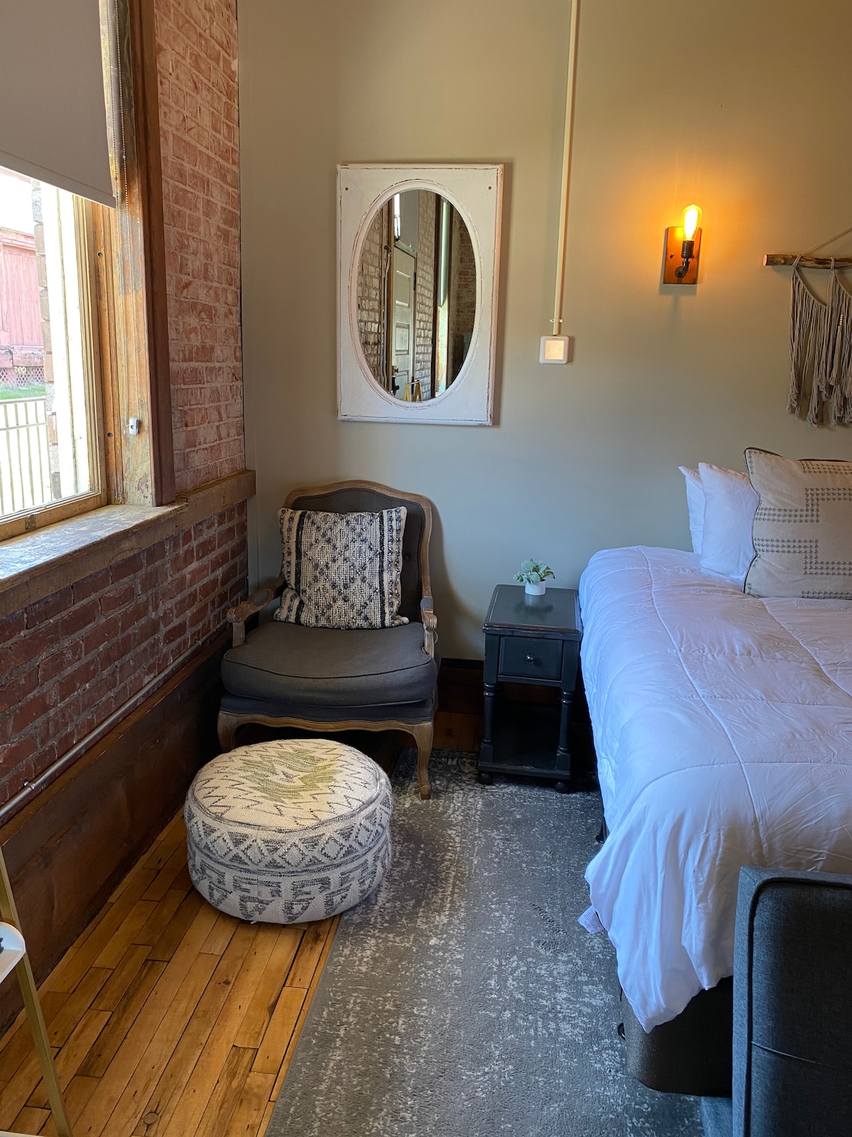 A cozy guest room features exposed brick walls and large windows allowing natural light. A spacious bed is complemented by a bedside table and a comfortable chair. An accent pouf and a decorative plant add subtle charm to the space.