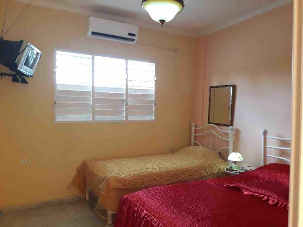 A well-lit bedroom features two beds, one covered with a red bedspread and the other with a gold one. A small nightstand with a lamp is positioned beside the beds. A wall-mounted television is visible, and natural light enters through a pair of shuttered windows.