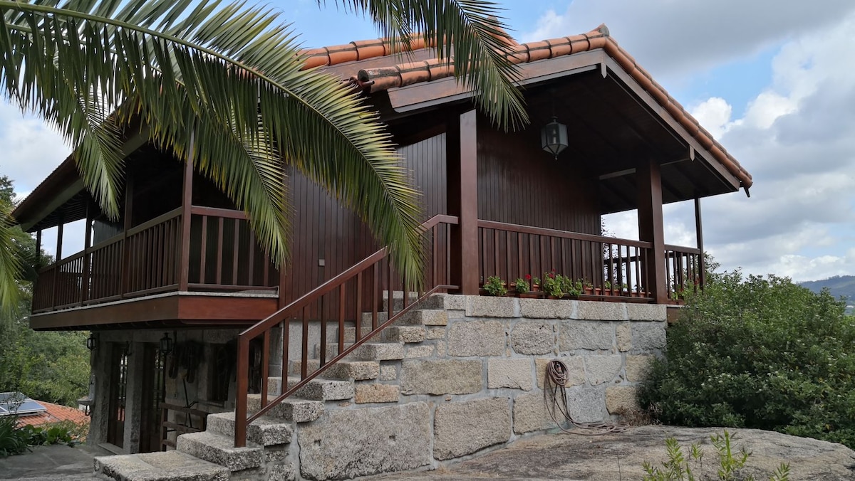 A rustic wooden house features a sloped roof and an elevated porch with railings. Stone steps lead up to the entrance, surrounded by lush greenery and palm leaves. The structure blends harmoniously with the natural landscape, creating a serene country retreat.