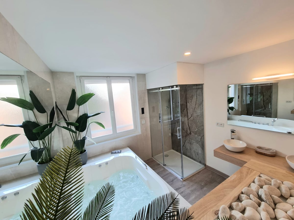 The spacious bathroom features a luxurious two-person balneo tub, surrounded by natural light from a nearby window. A modern glass shower is visible, along with a sleek wooden countertop hosting elegant sinks. Green plants add a touch of nature to the contemporary design.