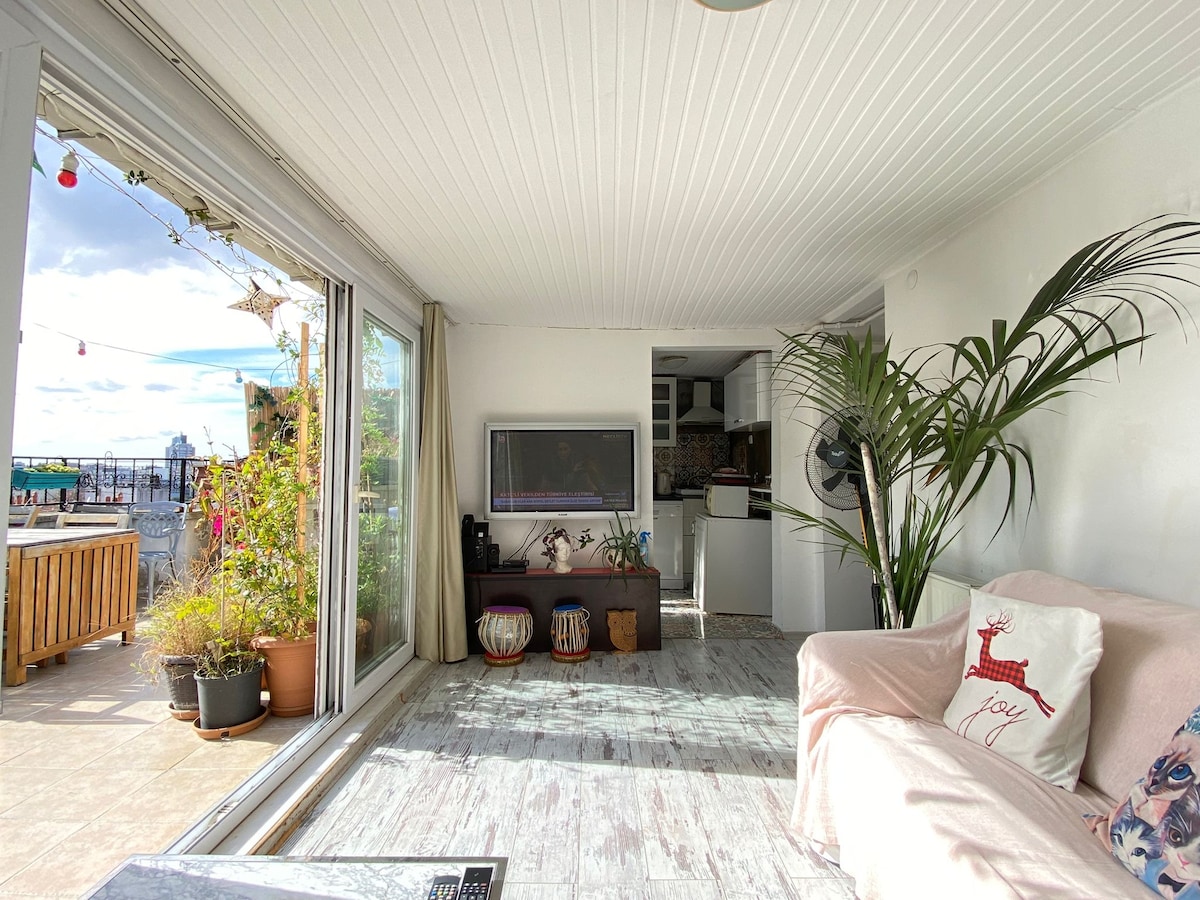 A light-filled living area features large glass doors that open to the terrace. A comfortable sofa is positioned beside a flat-screen television. Potted plants add greenery, while a wooden table complements the cozy atmosphere. The space is designed for relaxation and socializing.