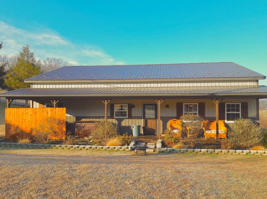 The exterior of Pond Hill Guesthouse is presented with a welcoming front porch, featuring seating areas and garden beds. A sturdy metal roof covers the structure, and a wooden fence adds privacy. The surrounding landscape includes a grassy yard with seasonal greenery.