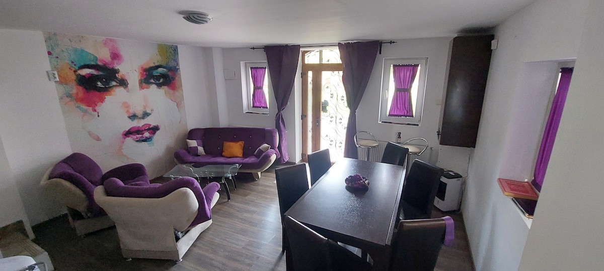 A modern living area features a striking mural on one wall, complemented by a purple sofa set and a dark wooden dining table. Natural light enters through the large window, framed by purple curtains. A small decorative bowl is centered on the table.