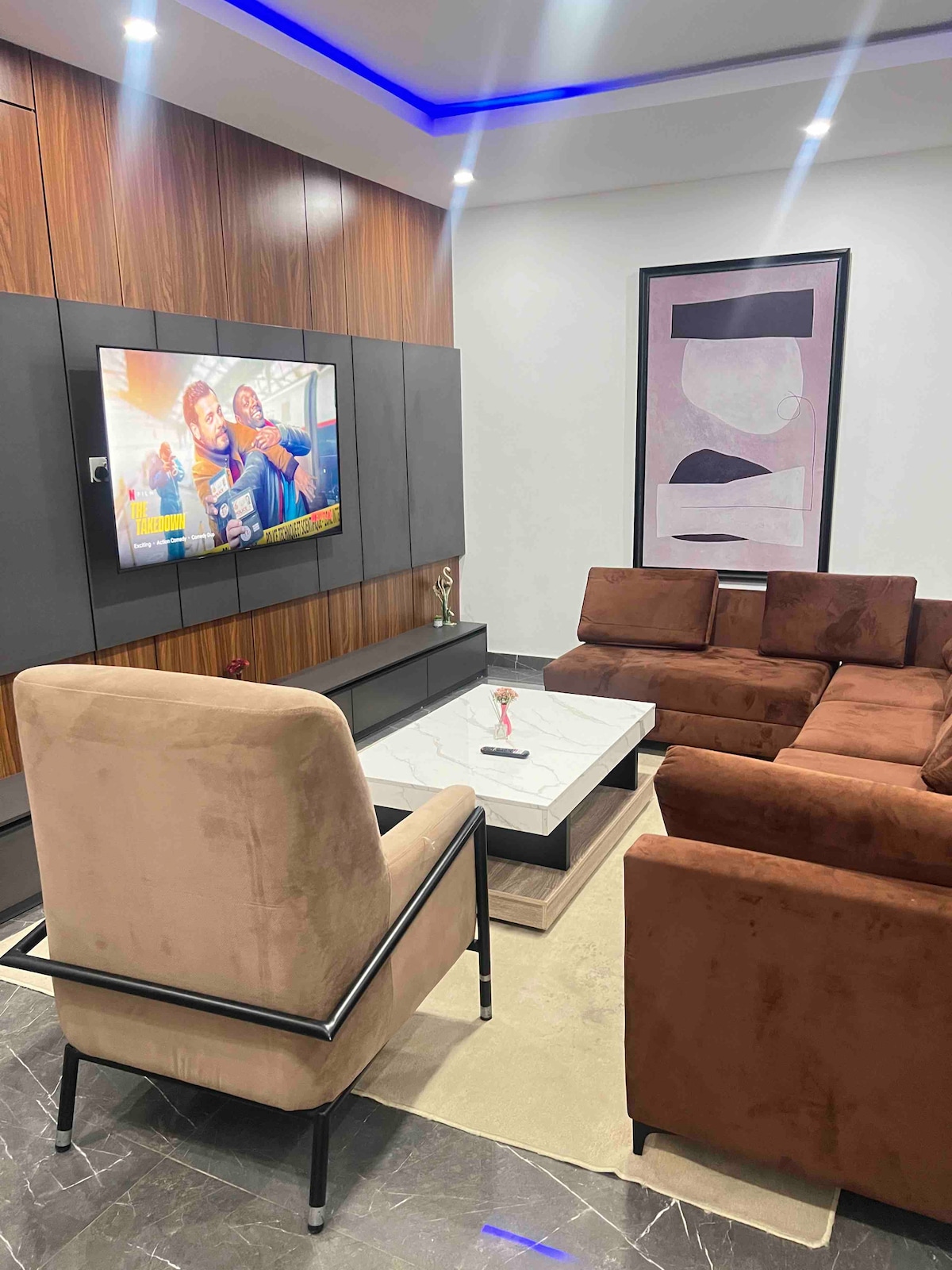 A modern living area features a sectional brown couch arranged around a light-colored coffee table. A plush armchair stands nearby, and a large flat-screen television is mounted on the wall, surrounded by stylish cabinetry. Soft lighting is provided by recessed fixtures above.