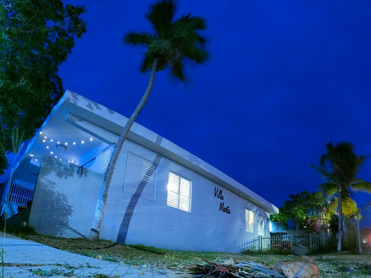A modern single-story apartment is viewed from an angle under a twilight sky, showcasing palm trees swaying nearby. Soft lighting illuminates the entrance, while the name 'Villa Nauta' is displayed on the exterior wall. A calm ambiance is created by the surrounding greenery.