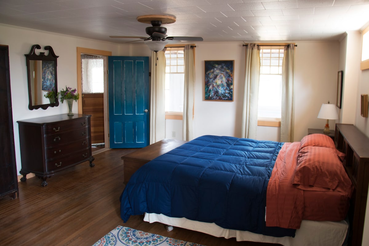 A spacious bedroom features a king-size bed with a blue quilt and rust-colored accent pillows. Two artworks adorn the walls, complemented by natural light from nearby windows. A decorative rug is placed under the bed, while a dresser and a mirror complete the inviting space.