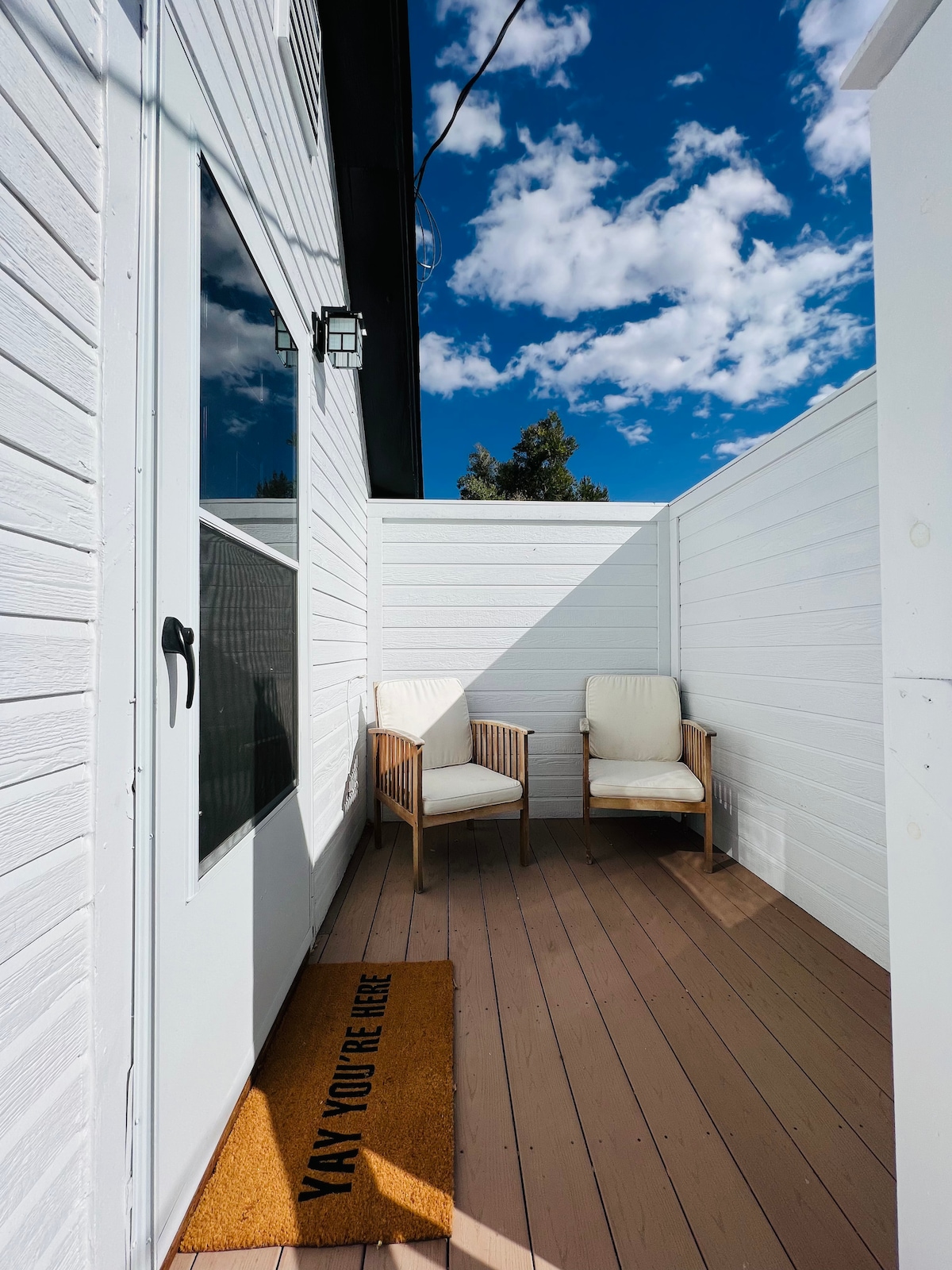 A private outdoor seating area features two wooden chairs with white cushions on a wooden deck. A welcoming doormat is placed at the entrance, and white walls create an enclosed space. The sky above is filled with fluffy clouds.