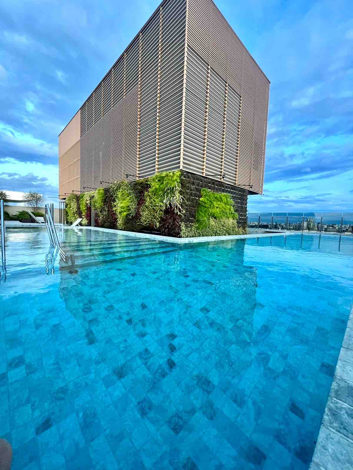 A rooftop pool is visible, featuring clear, tranquil water reflecting the blue sky. Surrounding greenery adds a touch of nature, while the modern building structure is partially seen in the background, presenting a clean, contemporary design.