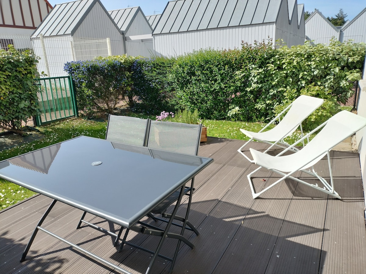 A private terrace features a grey table accompanied by two folding chairs. A pair of white lounge chairs are positioned nearby, inviting relaxation. Lush greenery and flowering plants create a natural border, enhancing the outdoor space.