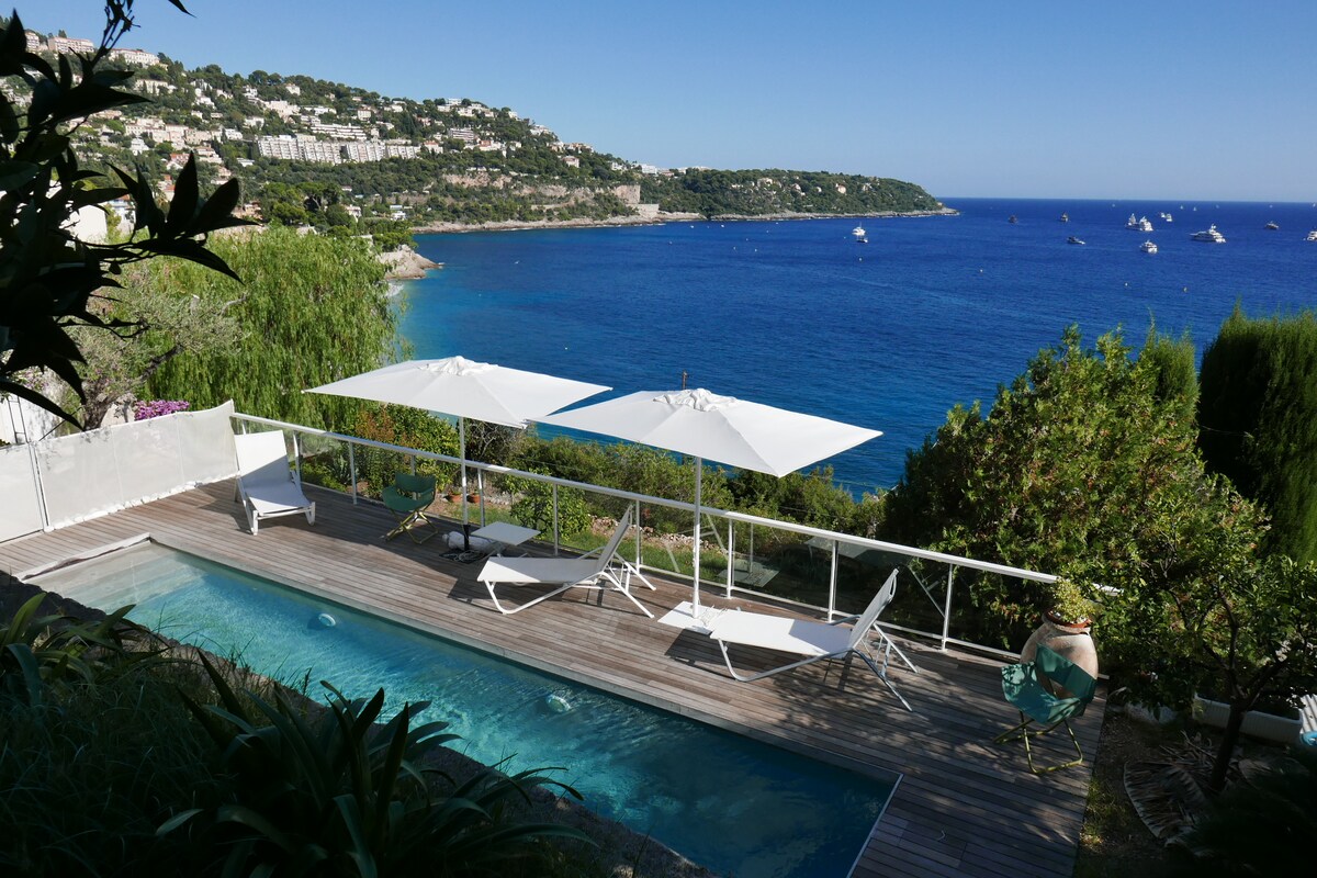 A private pool surrounded by wooden decking features two white umbrellas and lounge chairs positioned for relaxation. The sparkling blue waters of the sea are visible in the background, with lush greenery framing the scene and hills in the distance.
