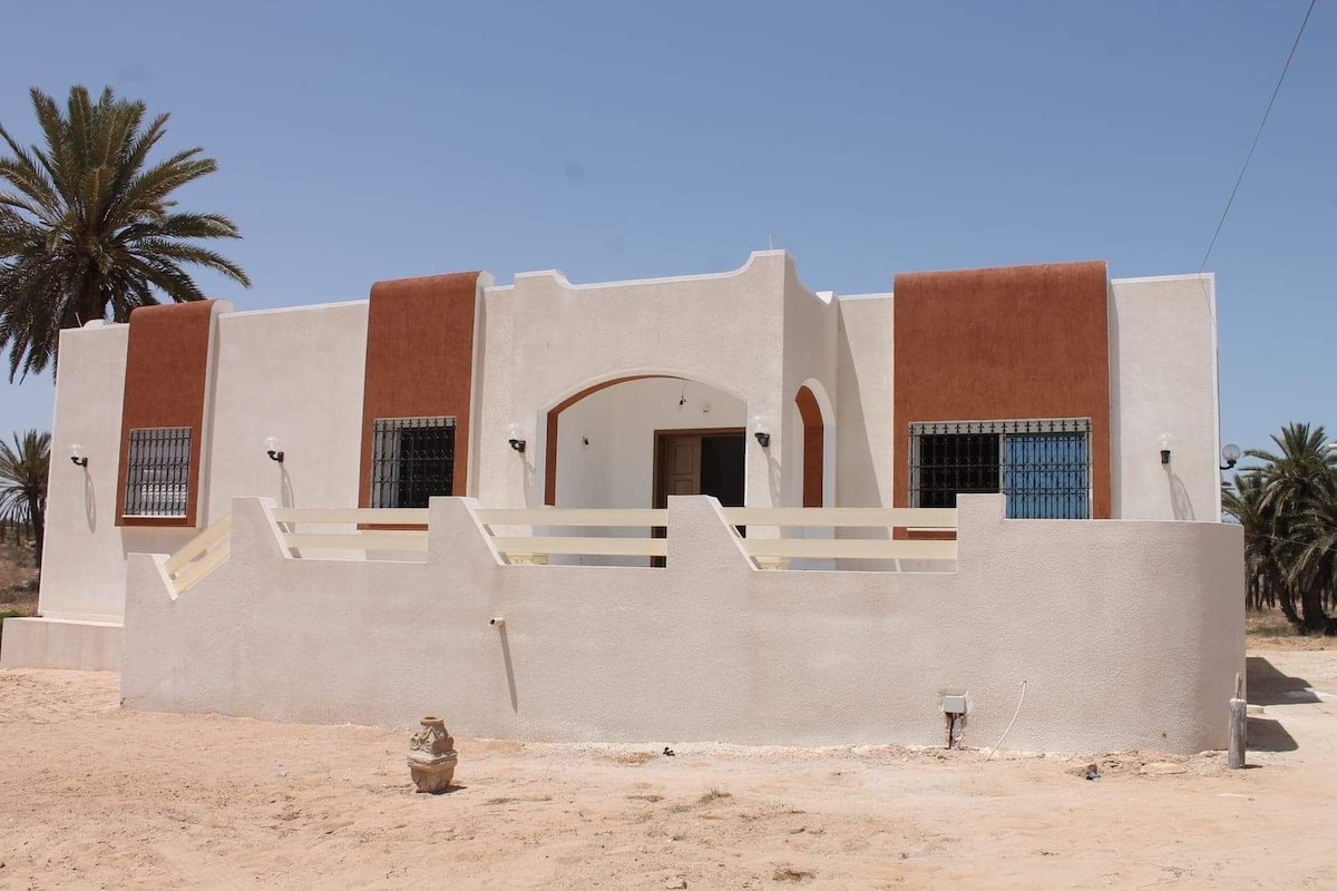 The exterior of the villa presents a modern design, featuring a smooth white facade complemented by earthy brown accents around the windows and entryway. Palm trees can be seen nearby, and the surrounding sandy area adds to the villa's tranquil setting.