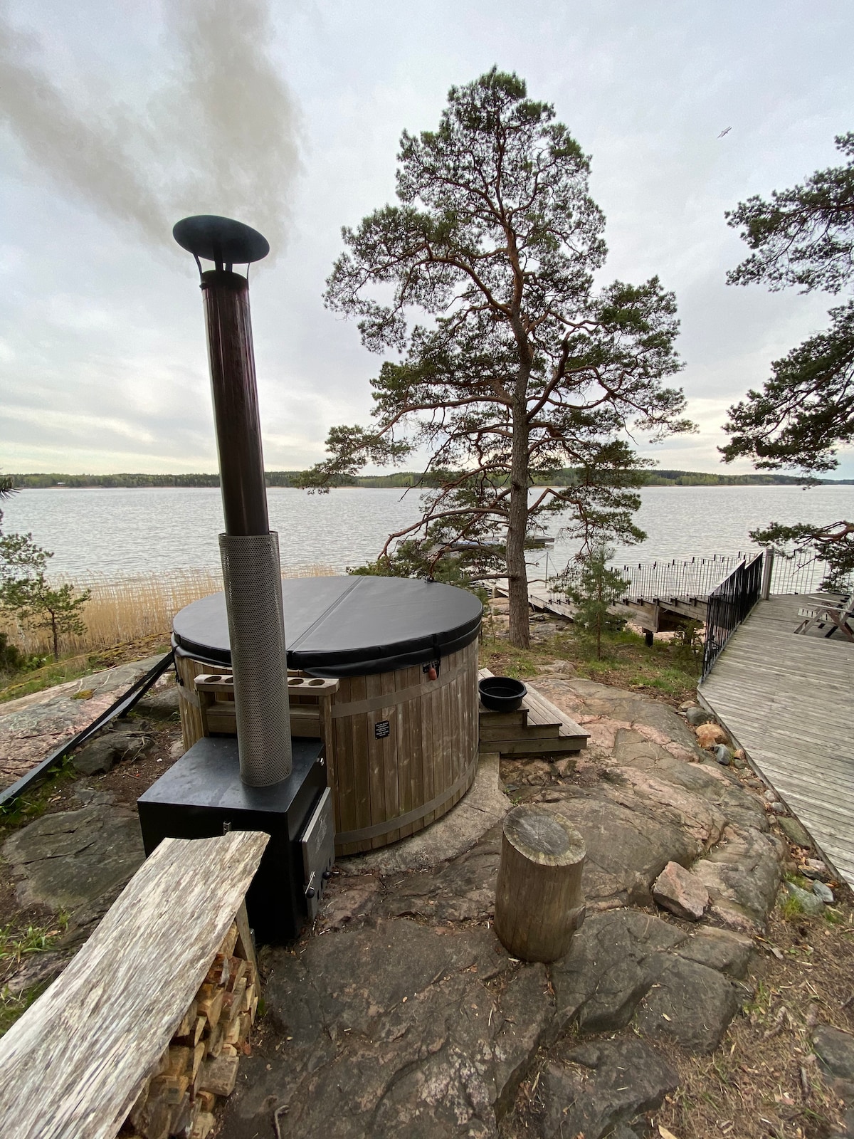 A wood-fired hot tub is situated on stone with scenic views of the water. A chimney extends from the hot tub, emitting steam. A tree stands nearby, enhancing the natural setting, while wooden pathways lead towards the water's edge.