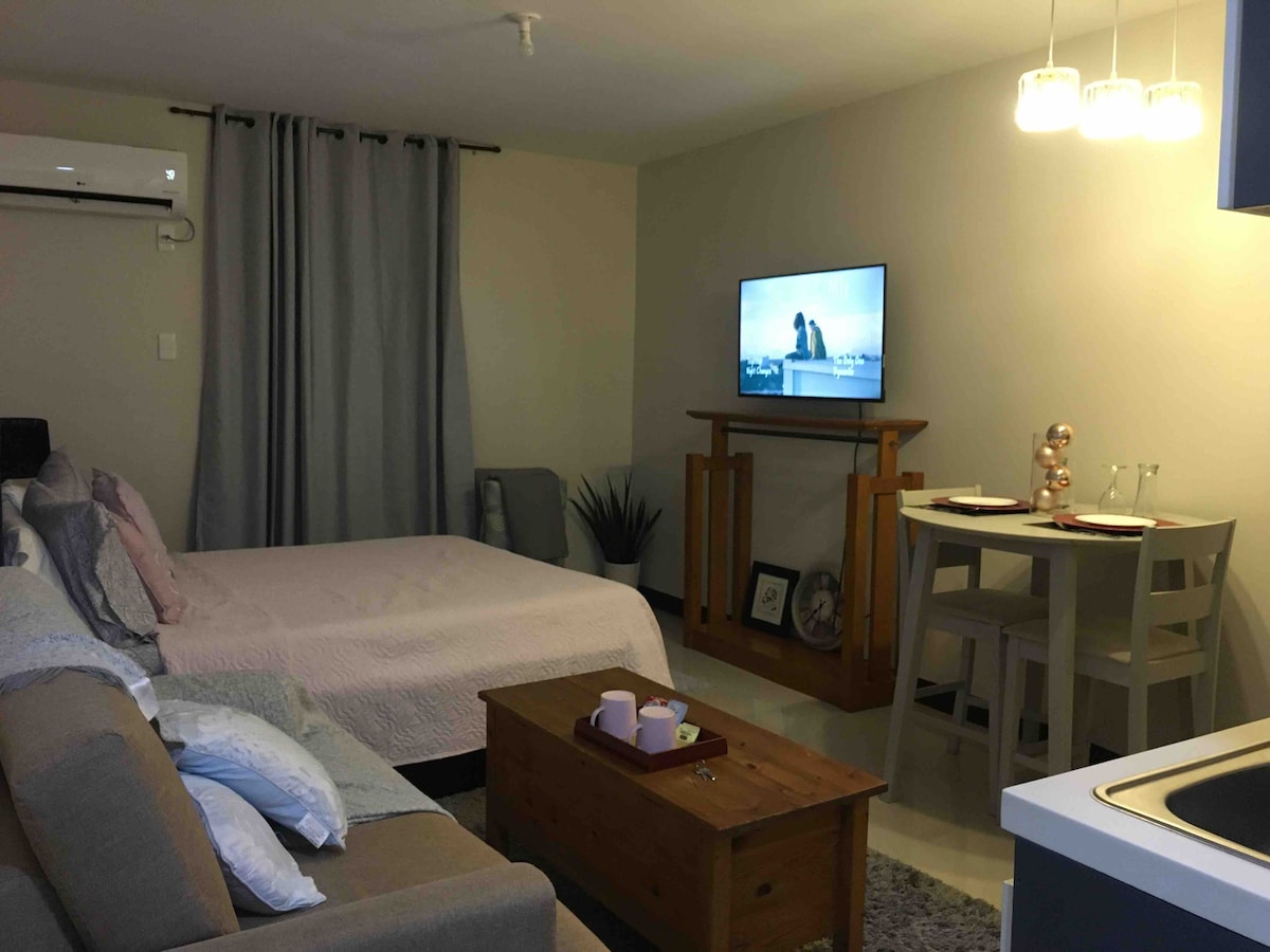 A cozy studio room is captured, featuring a spacious bed covered with a soft pink bedspread. A smart TV is mounted on the wall opposite a comfortable seating area and a wood coffee table. The dining space includes two stools at a compact table, with soft lighting overhead.