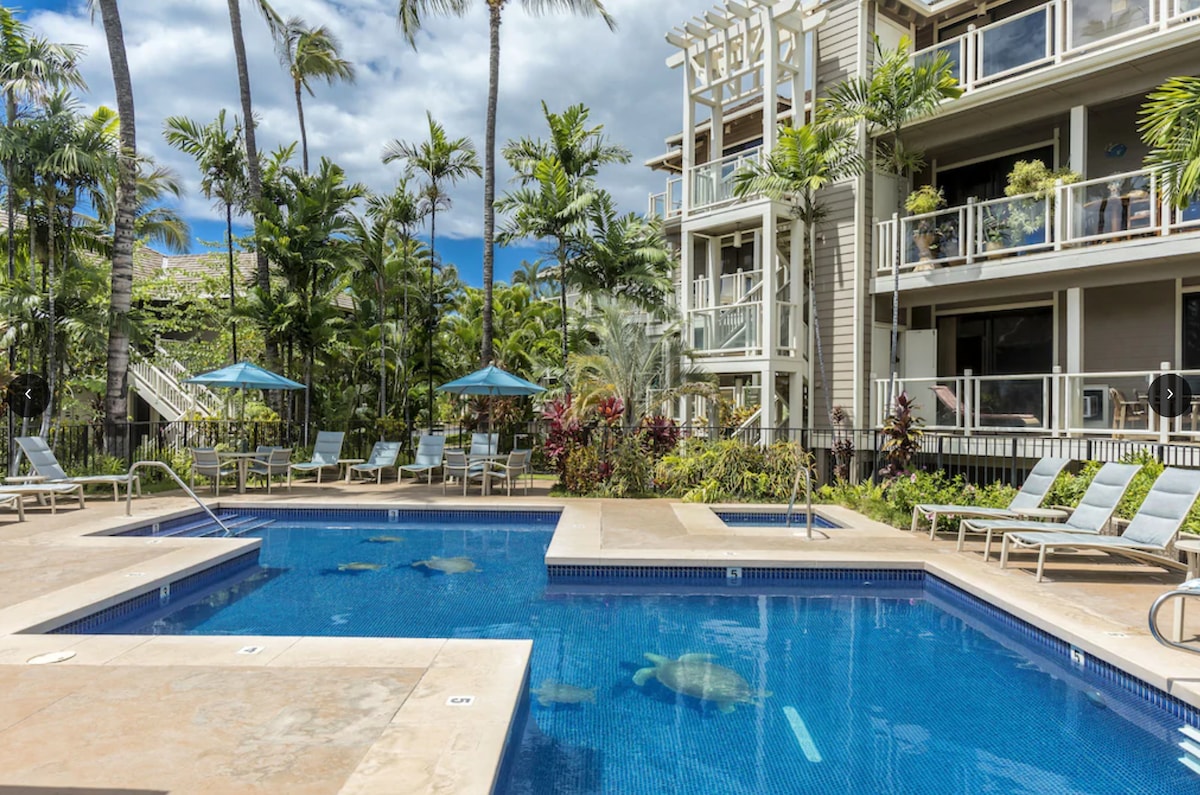 A beautifully maintained swimming pool is surrounded by lush tropical landscaping and palm trees. Lounge chairs with umbrellas are positioned near the pool, inviting relaxation. The patio of the nearby building offers an inviting view of the pool, creating a serene ambiance.