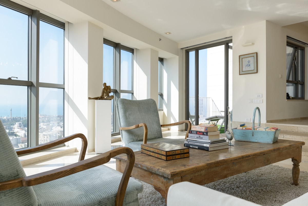 A bright living area features large windows offering panoramic views of the coast. Two comfortable armchairs are positioned around a wooden coffee table, topped with a stack of books and a small basket. Natural light enhances the spacious, welcoming environment.