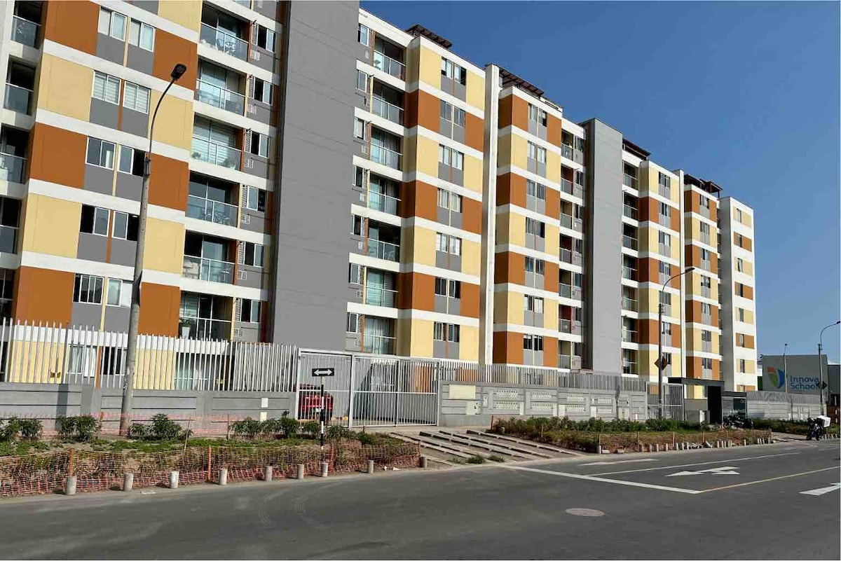A modern multi-story building is captured, showcasing a combination of yellow, orange, and gray exterior panels. The structure is lined with large windows, and a fenced area with landscaping features is visible in the foreground. Clear skies create a bright backdrop.