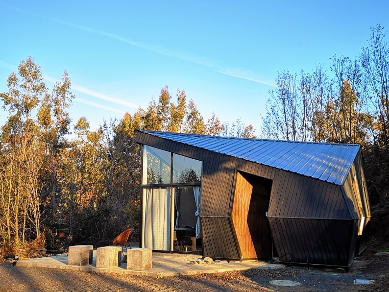 A modern cabin with an angled roof features large glass sections that invite ample natural light. Surrounded by trees, the structure showcases a combination of wooden and metal elements, with a fire pit area visible in front, creating an inviting outdoor space.