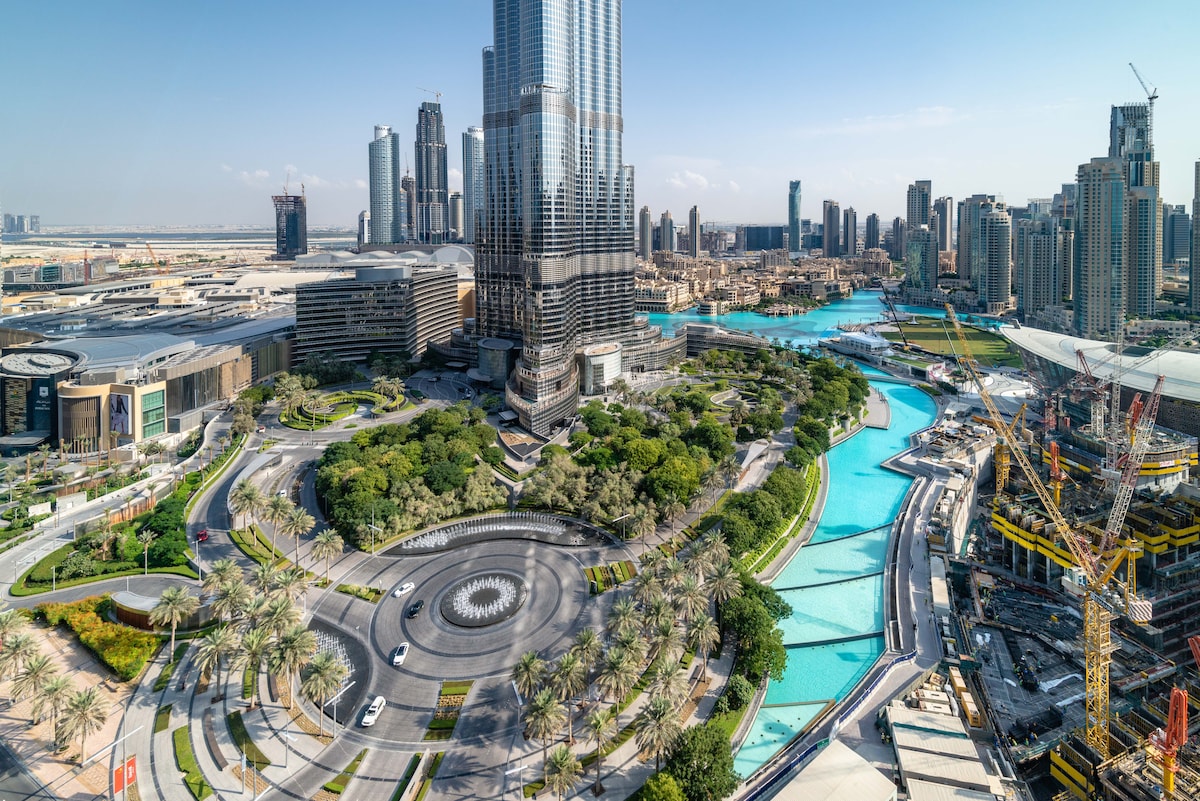 An aerial view captures the Burj Khalifa rising above a landscaped area with greenery and a turquoise waterway. The surrounding city showcases modern architecture, while vehicles navigate the circular road lined with palm trees. Construction activity is visible in the background.