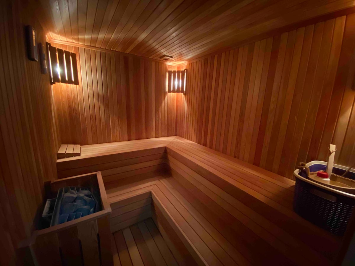 A warm sauna room is presented with wooden paneling covering the walls and seating areas. Soft lighting from wall fixtures enhances the natural wood tones, creating a serene environment. A wooden bucket and ladle are visible, providing tools for a traditional sauna experience.