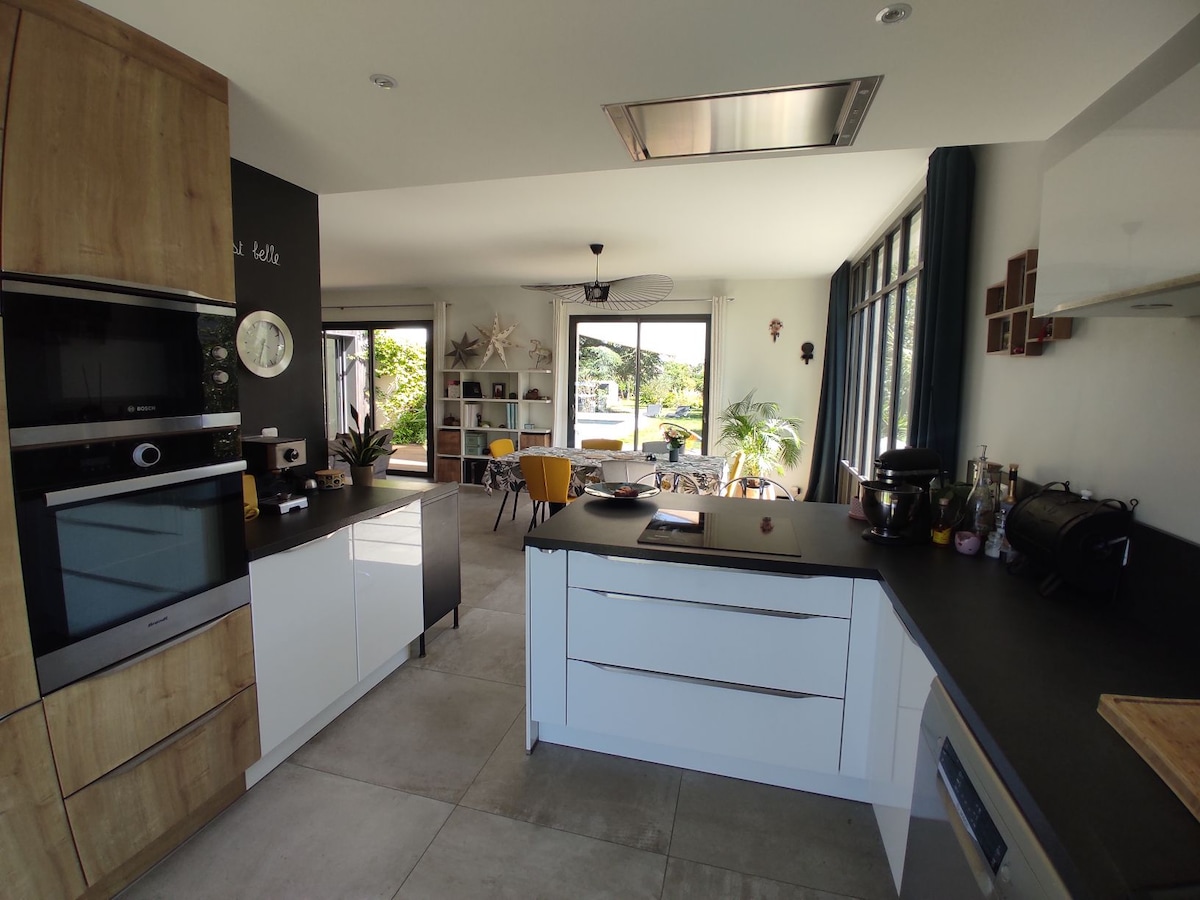 The kitchen features a combination of wood and sleek cabinetry, equipped with modern appliances and a central island. A spacious dining area is visible through large glass doors, allowing natural light to illuminate the open layout. Potted plants add a touch of greenery to the space.