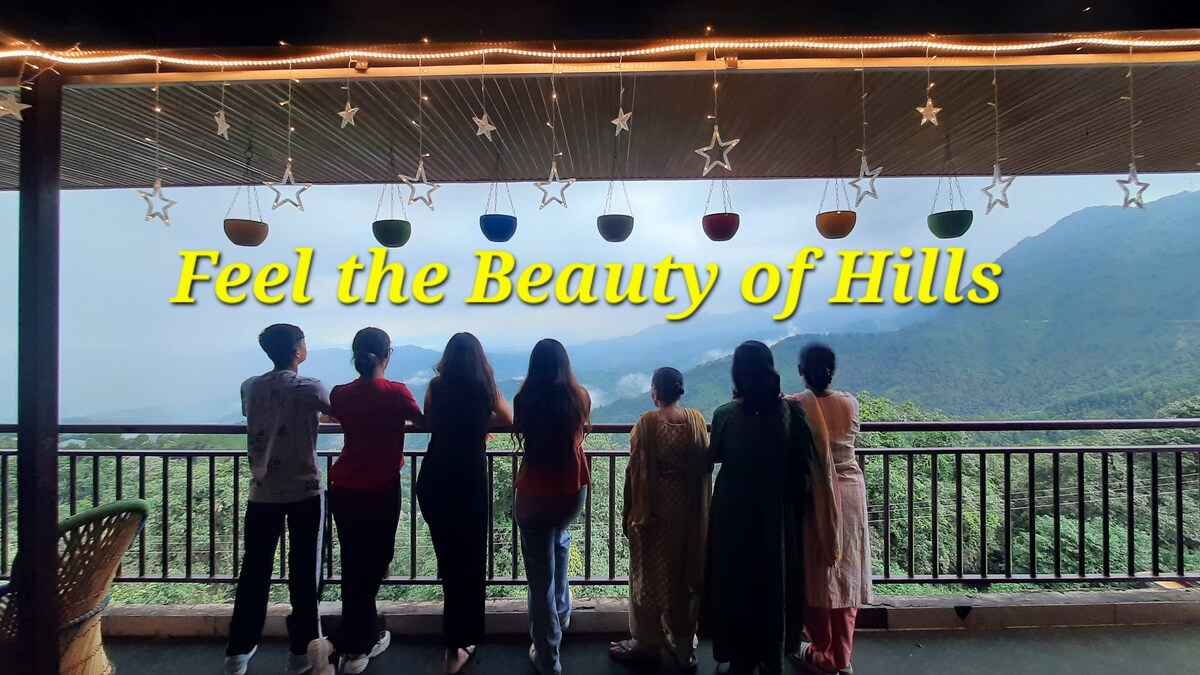 A group of individuals stands along a balcony railing, gazing at the misty hills below. Colorful decorative bowls hang from the ceiling, adding a touch of charm. The expansive view showcases lush greenery and a cloudy sky, enhancing the serene atmosphere.