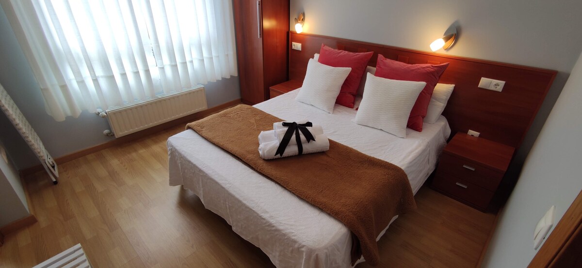 A well-lit bedroom features a spacious bed with white linens and decorative pillows in shades of red. A warm throw blanket is neatly arranged at the foot of the bed. Natural light enters through sheer curtains, enhancing the room's inviting aesthetic.