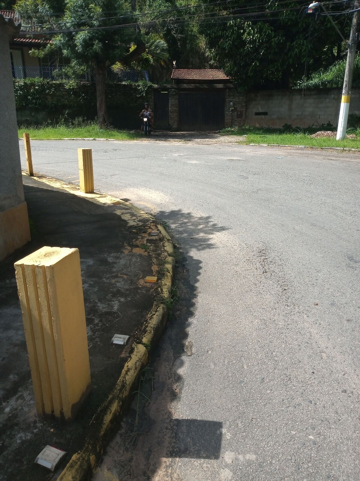 A quiet street corner is depicted, featuring yellow traffic posts along the curb. The asphalt road curves gently, bordered by greenery and a glimpse of a wooden gate in the background. Scattered foliage complements the serene environment.