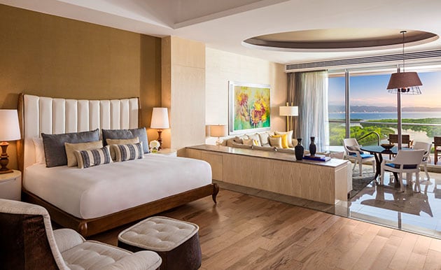 A spacious living area features a king-size bed, modern art, and a cozy seating arrangement. Large glass doors lead to a private terrace, offering scenic views. Soft hues and natural light create a peaceful atmosphere, while polished wood flooring adds warmth.