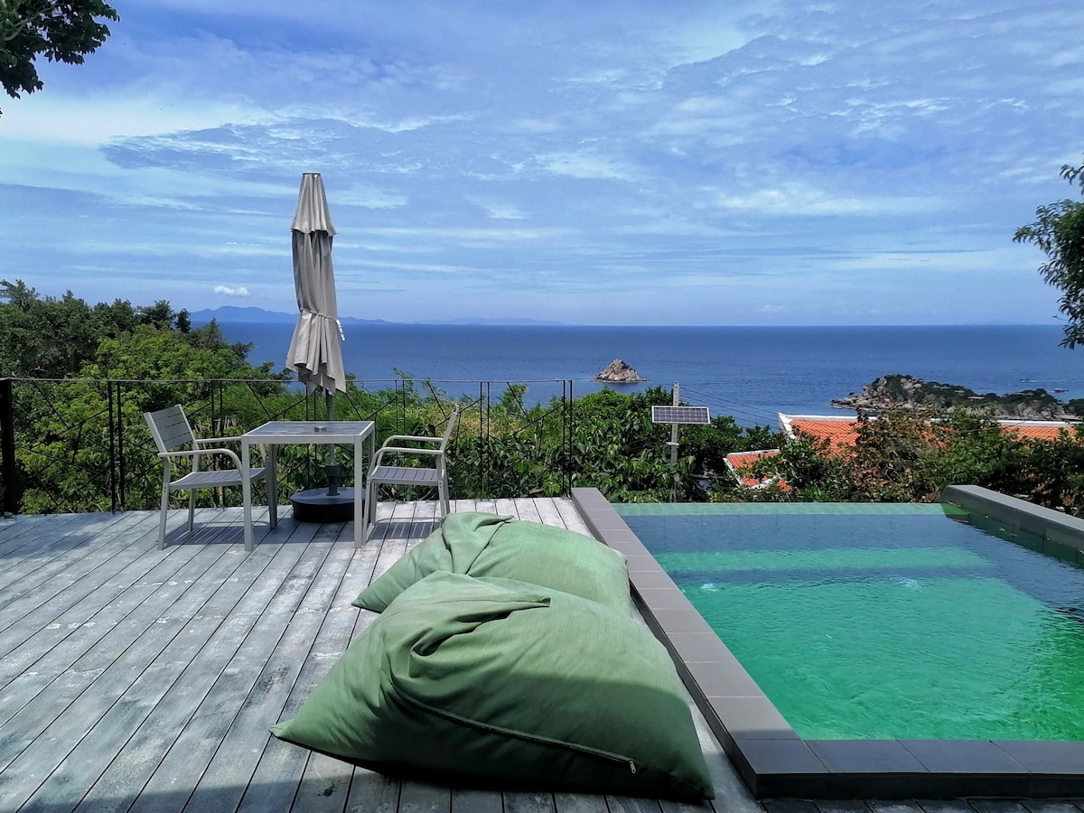 An outdoor deck features a private pool with clear water reflecting the sky. A dining table and chairs are positioned nearby, while a large umbrella provides shade. Lush greenery and an ocean view add to the serene setting.
