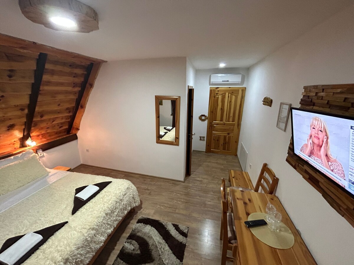 A well-lit room features a spacious bed with soft bedding, complemented by wooden accents on the walls. A small dining table with chairs sits beside the bed, and a flat-screen TV is mounted on the wall. A mirror reflects the inviting space.