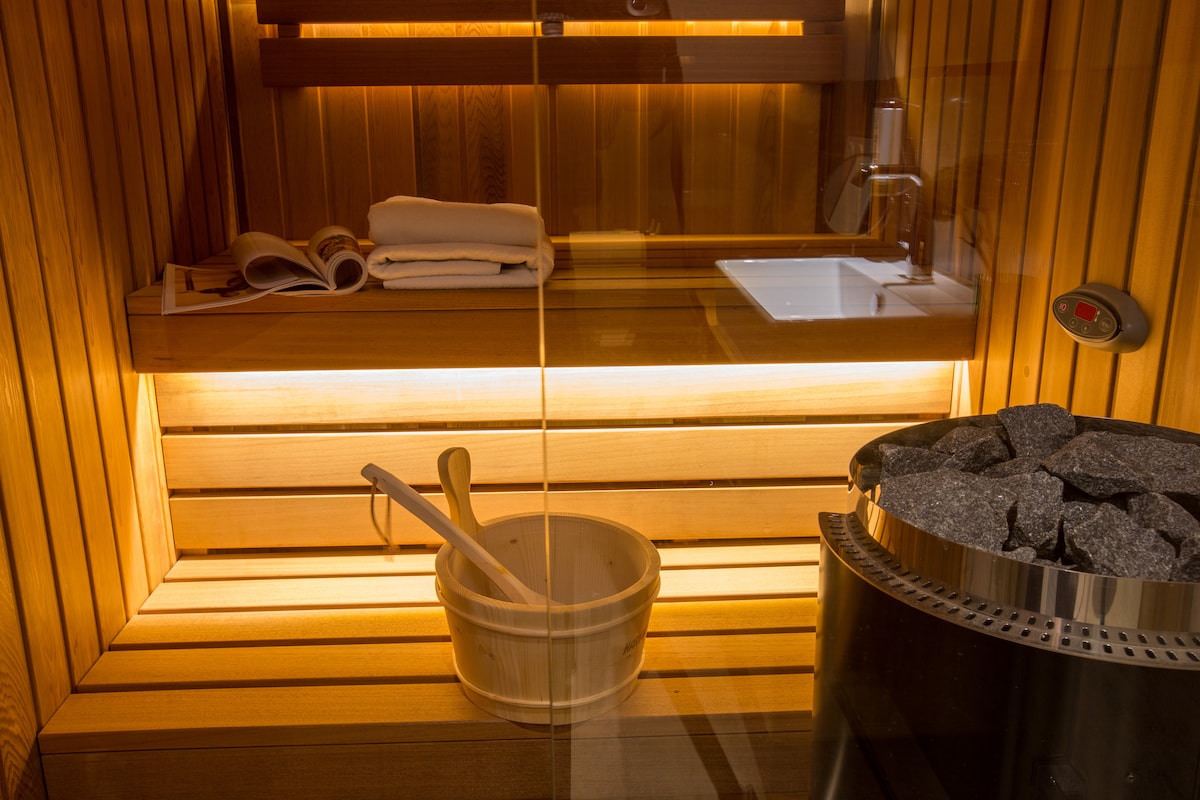 A wooden sauna interior is illuminated with warm lighting. The space includes smooth wooden benches, a stone heater filled with stones, a wooden bucket, and a ladle. Towels are neatly arranged on a shelf, enhancing the inviting atmosphere of relaxation.