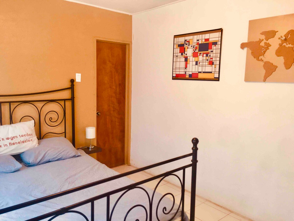 A cozy bedroom features a black metal bed with a blue bedspread and decorative pillow. A bedside table with a lamp is positioned beside the bed. A wooden door allows access to the adjoining area. Wall art, including a world map and a colorful grid, adorns the walls.