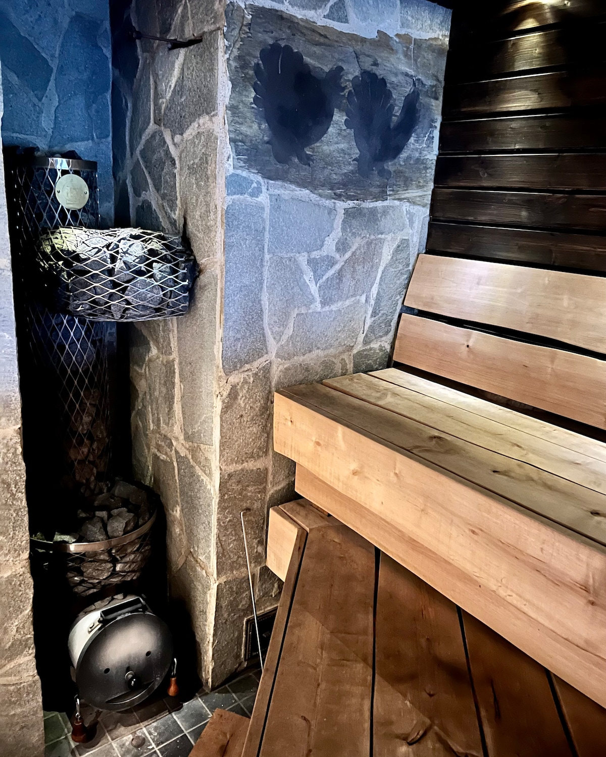The image displays the interior of a traditional wood-heated sauna. Wooden benches are positioned along the walls, complemented by stone accents. A stone stove and a container of stones are visible, providing warmth. The wall features decorative handprints, adding a unique touch to the space.