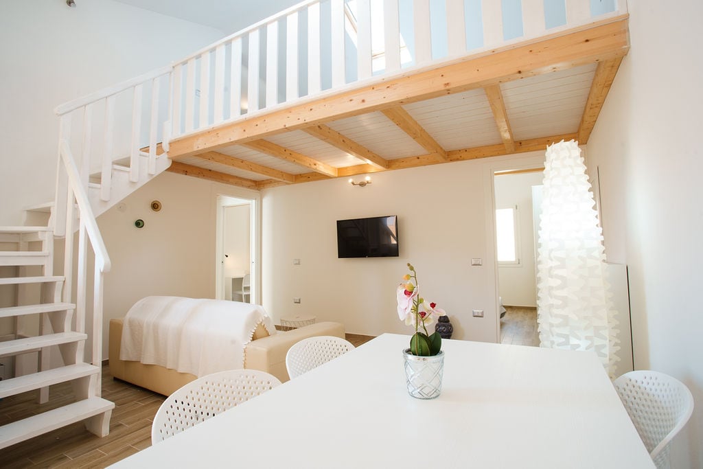 A bright living area features wooden beams and an open loft space. A white dining table is surrounded by modern chairs, with a small potted plant at its center. A comfortable sofa is positioned nearby, and a television is mounted on the wall, enhancing the inviting atmosphere.
