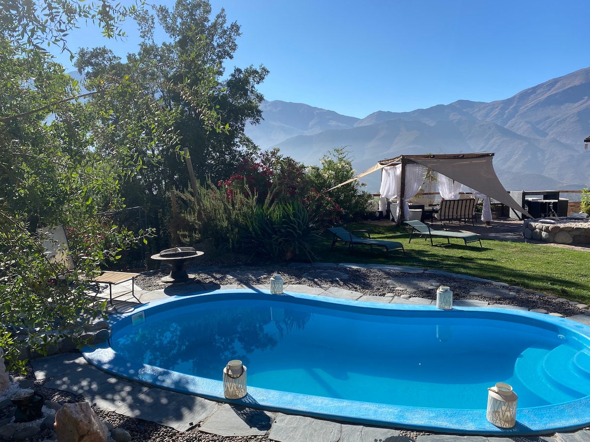 A blue swimming pool is surrounded by greenery and mountain views. Nearby, lounge chairs invite relaxation, with a shaded seating area under a draped canopy. Lanterns line the pool, adding a gentle touch to the outdoor space, which enjoys abundant natural light.
