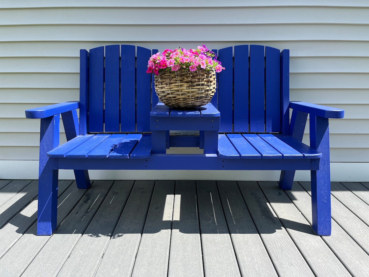 A vibrant blue bench is positioned on a wooden deck, featuring a central table between two seating areas. A wicker basket filled with colorful flowers rests atop the table, enhancing the inviting appearance of the outdoor space.