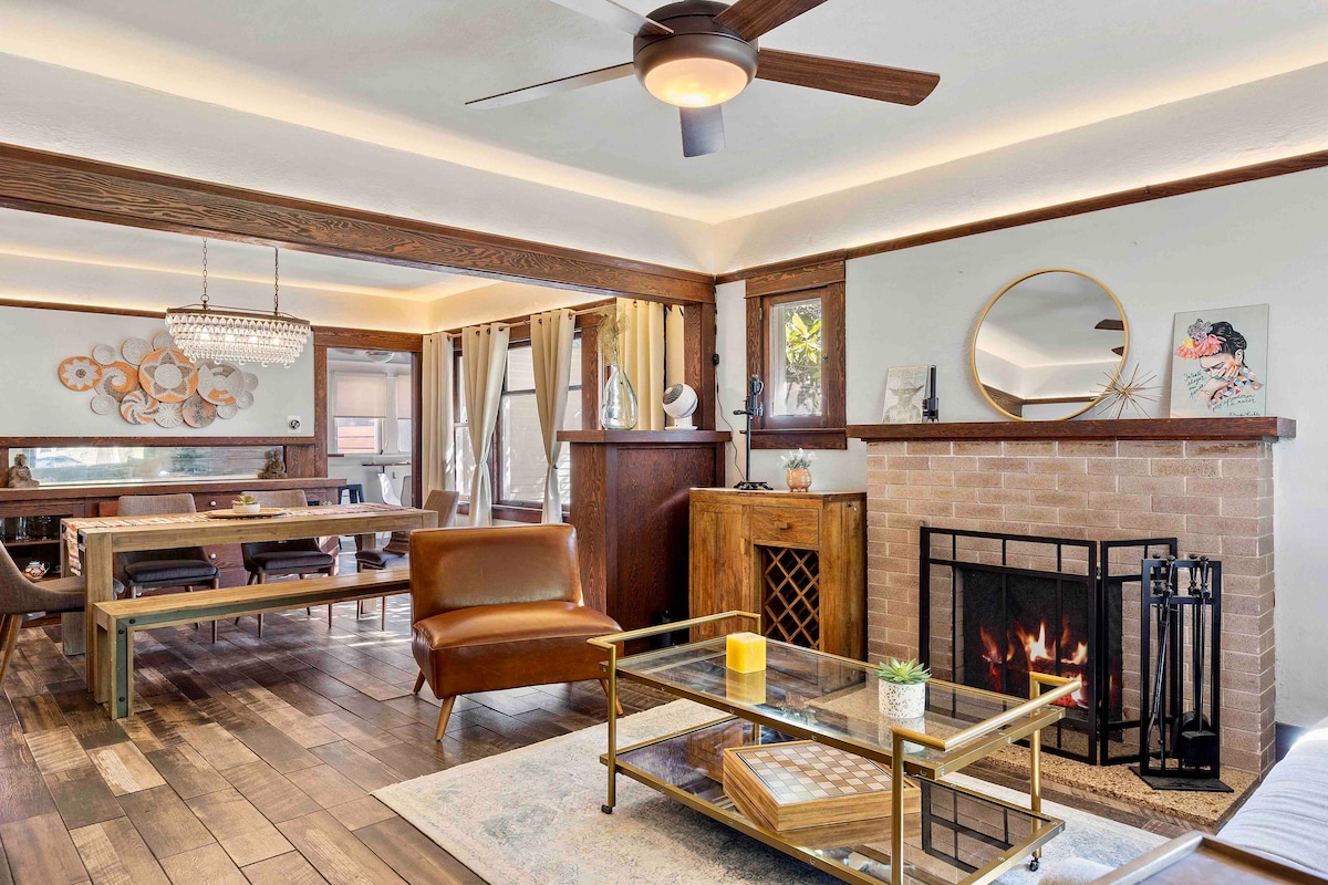 A cozy living space features a brown leather chair and a glass coffee table within view of a decorative fireplace. Large windows allow natural light to fill the room, while wood accents and a contemporary chandelier add character to the area.
