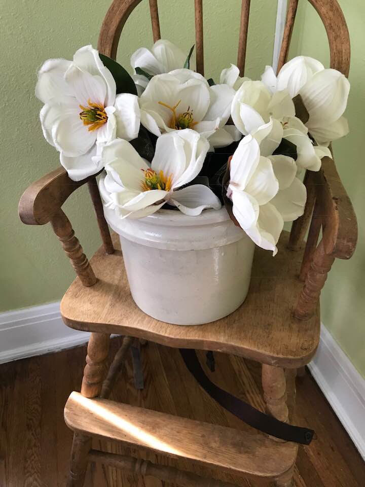 A rustic wooden chair sits against a light green wall, showcasing a white ceramic pot filled with large, artificial white flowers. The natural wood tones of the chair contrast with the vibrant blooms, creating a serene focal point.