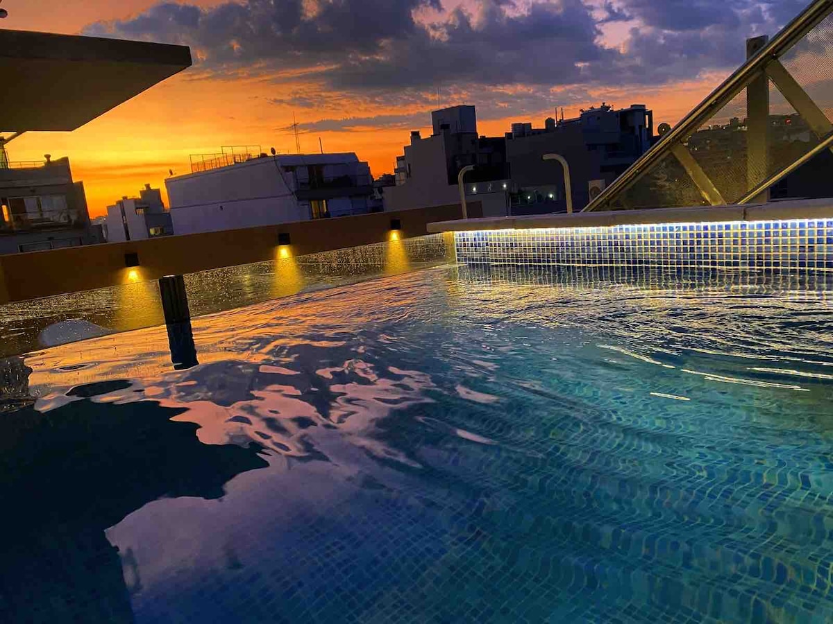 The rooftop pool reflects a vibrant sunset, with hues of orange and purple visible on the calm water's surface. Soft lighting fixtures line the pool's edge, creating a serene ambiance against the backdrop of urban architecture.