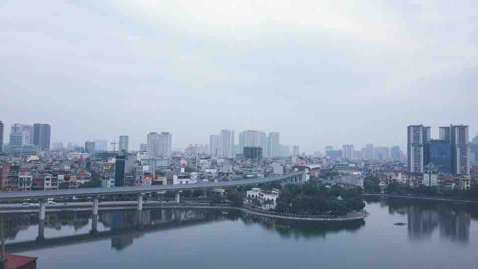 A view of the surrounding city is captured across a serene lake. Residential and commercial buildings rise in the background, with a bridge crossing over the water. The skyline displays a mix of modern architecture and greenery along the lake's edge.