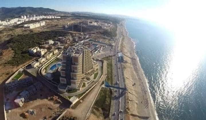 An aerial view captures a coastal landscape featuring a tall building overlooking the water. The beachfront stretches along the shoreline, and a road runs parallel. Rolling hills and distant urban development are visible in the background under a clear blue sky.