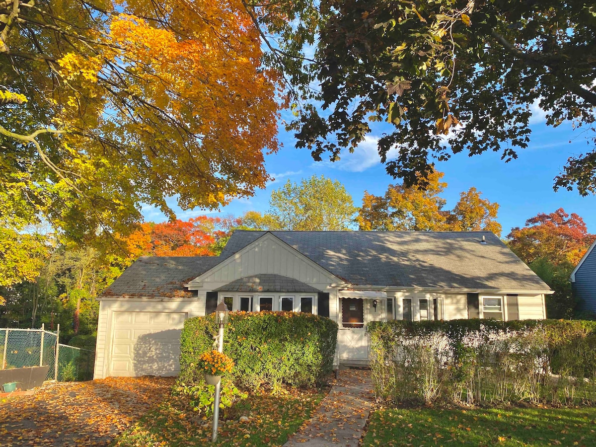 A single-family home is framed by trees with autumn foliage, showcasing vibrant orange and yellow leaves. A stone path leads to the entrance, which is complemented by a well-maintained front yard and shrubbery. Sunlight bathes the exterior, creating a warm ambiance.
