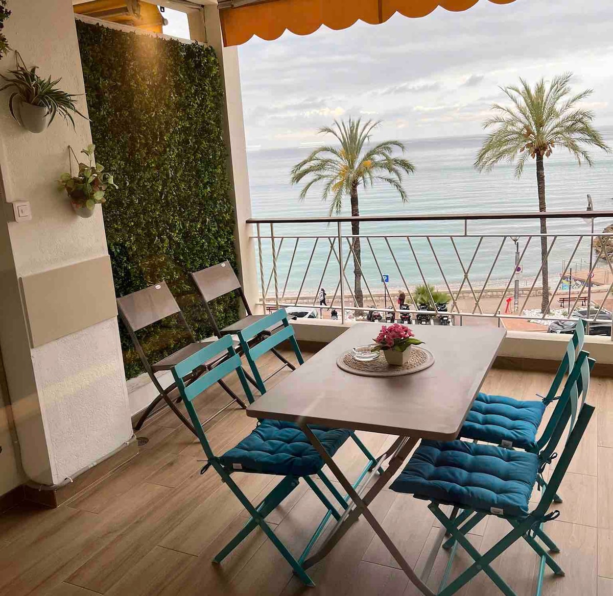 An inviting terrace features a table at its center surrounded by bright blue folding chairs. Lush greenery decorates one wall, while the ocean view is visible beyond the railing. Soft light filters through the overhang, creating a relaxed outdoor dining space.