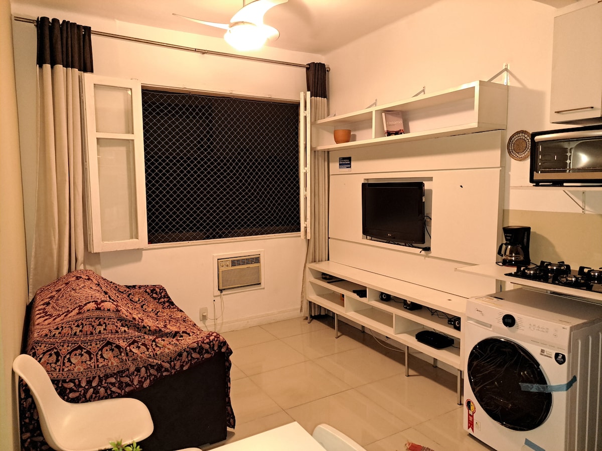 A cozy living area features a patterned throw draped over a comfortable couch. A wall-mounted television is positioned above a minimalist TV stand. Natural light enters through a window, and an air conditioning unit is visible beside it. A washing machine is integrated into the room's design.
