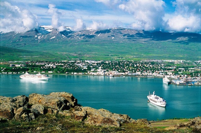 A panoramic view of a coastal town is presented, with ships docked in the harbor. Lush green hills and mountains create a scenic backdrop, while scattered clouds drift across a blue sky. The vibrant water reflects the landscape, enhancing the serene atmosphere.