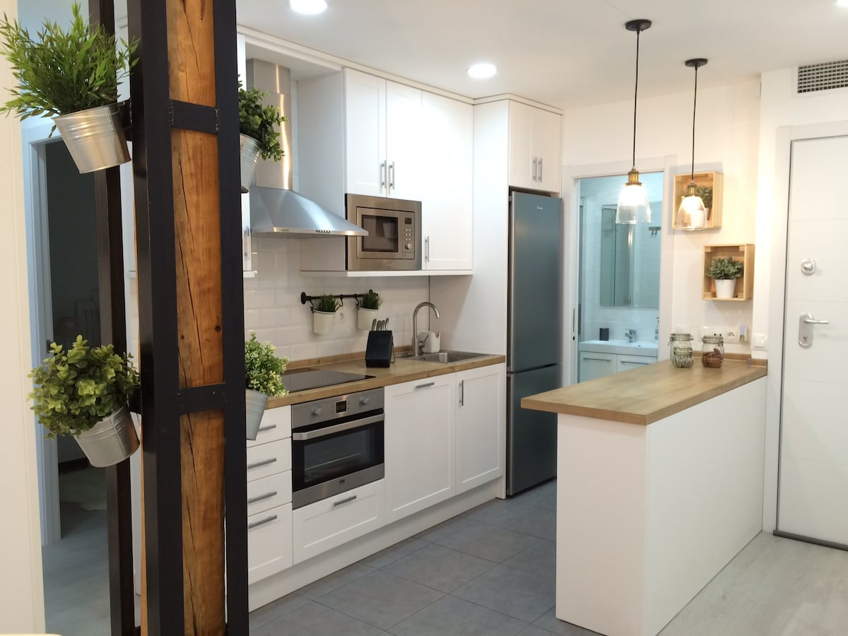 A modern kitchen features white cabinetry with wooden countertops and stainless steel appliances. Pendant lighting hangs above the kitchen island, and potted plants are arranged on a vertical structure. A window allows natural light to enter, illuminating the organized workspace.