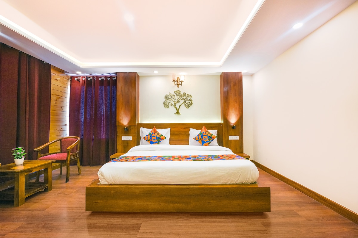 A spacious sleeping area features a wooden bed with colorful decorative pillows. Ambient lighting is provided by wall sconces beside the bed. A small table with a potted plant is positioned next to a wooden chair. Rich curtains frame a window, complementing the warm wooden tones.