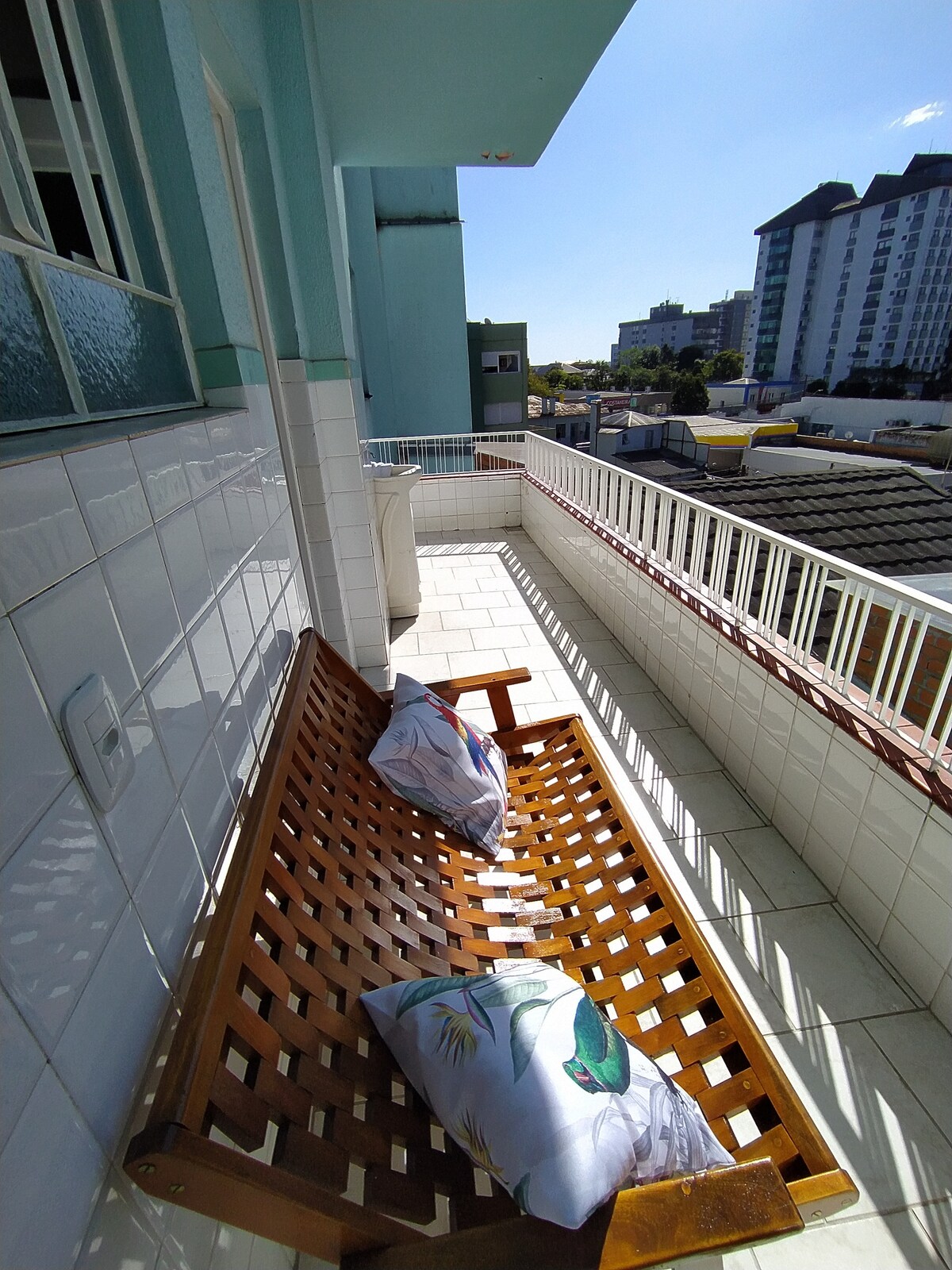 A wooden bench with decorative pillows is positioned on a balcony. The tiled floor extends outward, offering a view of the surrounding buildings and clear blue sky. A white railing encloses the space, enhancing the outdoor area.
