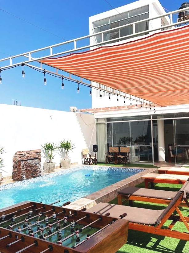An outdoor area featuring a pool with vibrant blue water, complemented by a wooden foosball table and two lounge chairs. Colorful striped awnings provide shade over the space, while potted plants add greenery. The inviting atmosphere is created by warm sunlight and a clear sky.