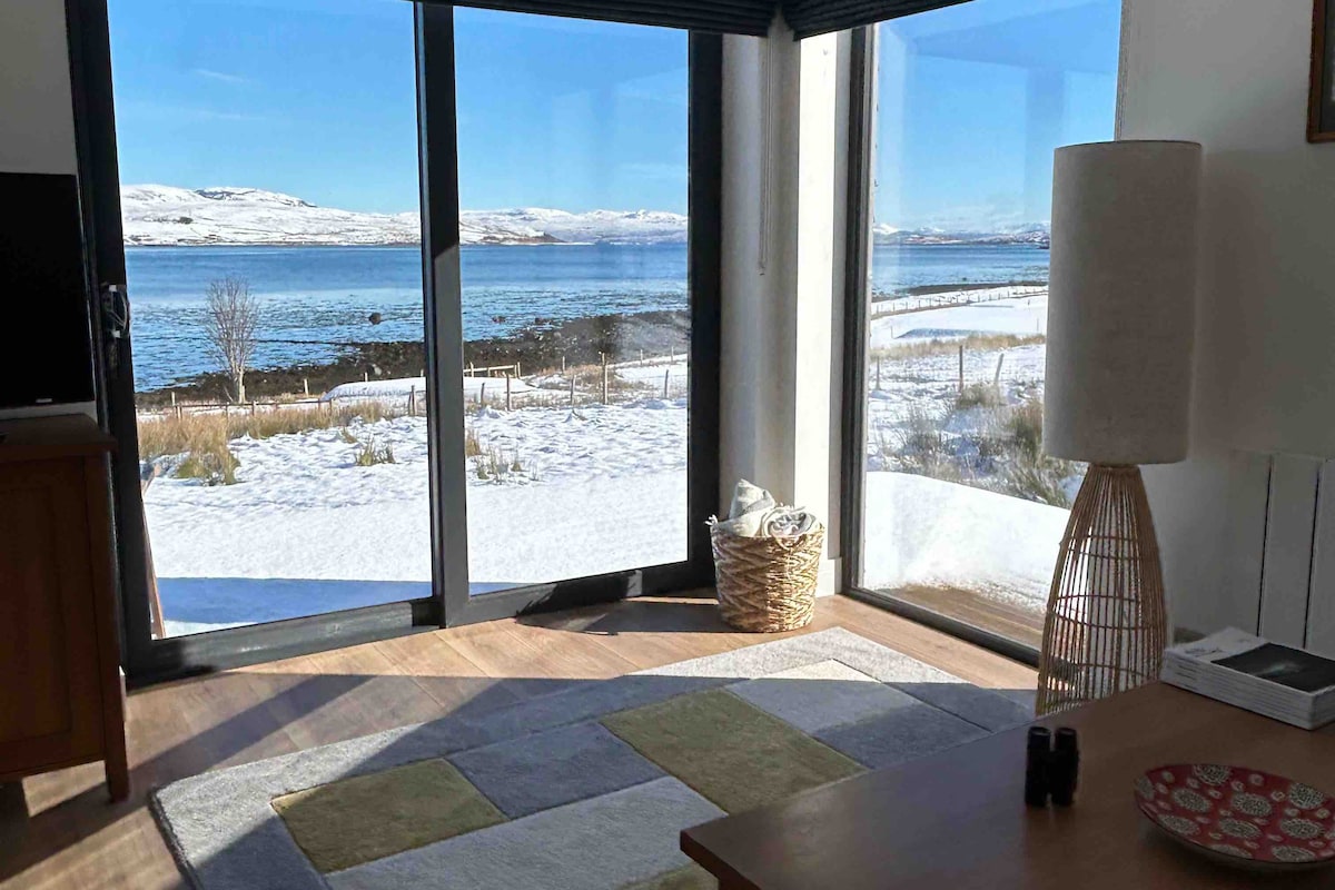 A corner seating area features large glass doors that open to showcase views of the water and snow-covered landscape. A soft area rug is placed on the wooden floor, while a woven basket and a floor lamp add functional decor to the space.
